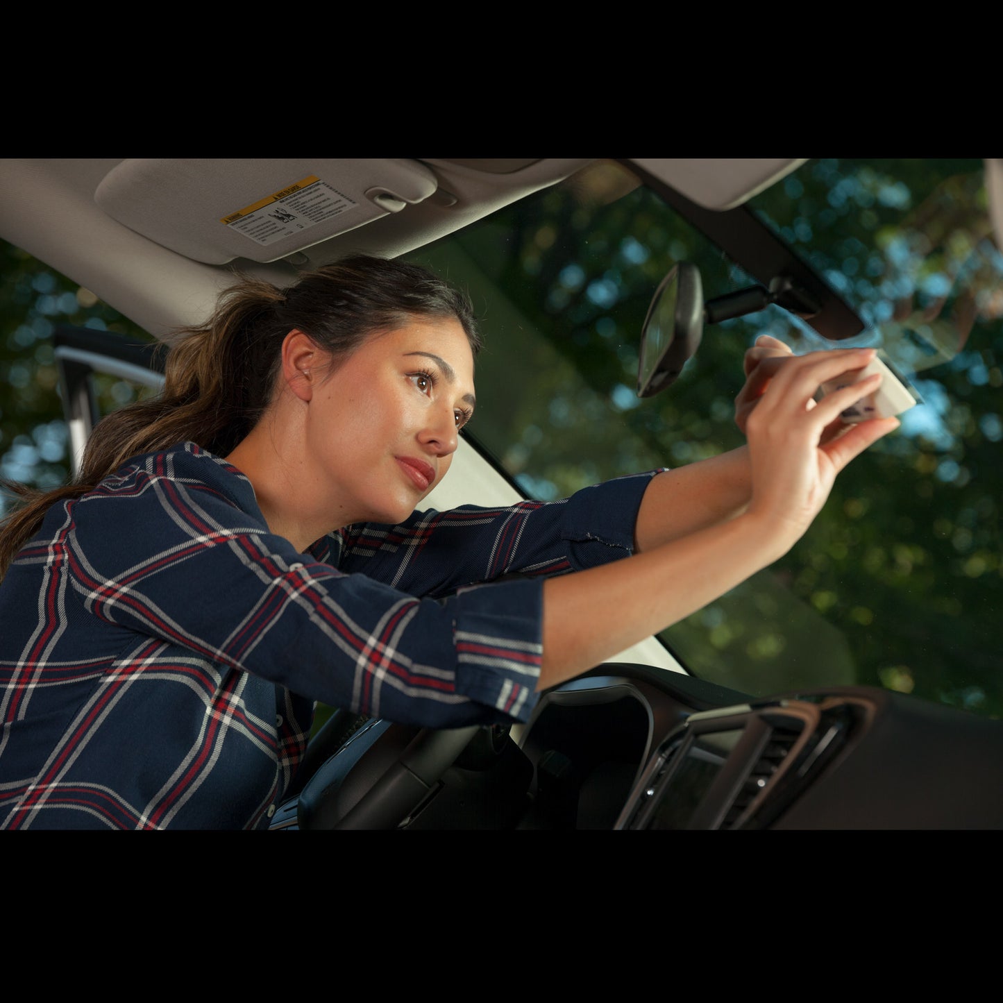 A woman gently pushes a toll transponder on to her windshield using sticker shield which is nearly invisible.